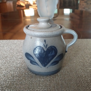 May include: A small, gray ceramic jar with a lid and a blue heart design. The jar has a handle and is decorated with a blue heart and leaves.