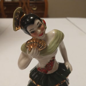 May include: A porcelain figurine of a woman in a flamenco dress. The figurine has black hair, red lips, and a gold fan. The dress is white with red floral accents and a dark green skirt with gold trim. The figurine is posed in a dance.