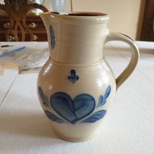 May include: A cream-colored ceramic pitcher with a blue heart design and blue floral accents. The pitcher has a wide mouth and a handle.