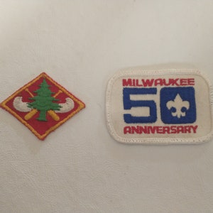 May include: Two embroidered patches. One is a red diamond with a green tree, two canoes, and two paddles. The other is a white rectangle with "MILWAUKEE 50 ANNIVERSARY" in blue and red, with a blue fleur-de-lis.