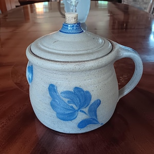 May include: A stoneware pottery jar with a lid and handle. The jar is light gray with a blue floral design and a blue rim. The lid has a cork stopper. The jar is sitting on a wooden table.