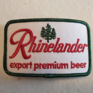 May include: Embroidered patch with the word "Rhinelander" in red script, a green tree graphic, and the text "export premium beer" on a white background. The patch has a green border.
