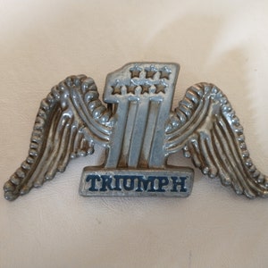 May include: Silver-colored metal pin with a winged number one and the word "TRIUMPH" in blue lettering.