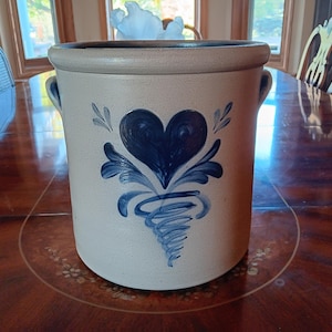 May include: A stoneware crock with a light beige base and a dark blue heart design. The crock has a cylindrical shape with two small handles and a dark rim. The heart design is surrounded by stylized leaves and a swirl. The crock is likely used for storage or decoration.