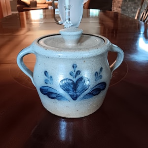 May include: A ceramic sugar bowl with a lid and two handles. The off-white bowl features a blue heart and floral design. The bowl is sitting on a dark brown wooden table.
