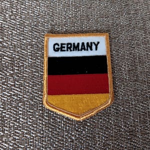 May include: Embroidered patch featuring the German flag design. The patch has a white top section with the word "GERMANY" in black, followed by black, red, and gold stripes. The patch has a gold border.