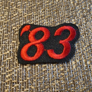 May include: Black rectangular patch with the red number "83" embroidered on it. The number is in a stylized font. The patch is on a textured, neutral-colored surface.