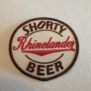 May include: A circular embroidered patch with a cream-colored background and a dark brown stitched border. The text SHORTY is stitched in brown at the top, Rhinelander in red cursive in the center, and BEER in brown at the bottom.