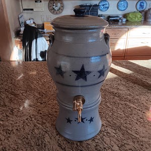 May include: A gray ceramic water dispenser with a spigot and a lid. The dispenser has a design of three black stars on the front.
