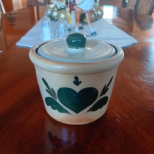 May include: A small, ceramic jar with a green heart design and a lid. The jar is decorated with green leaves and has a light brown glaze.