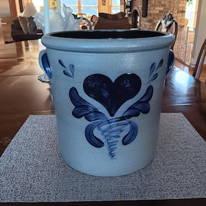 May include: A stoneware crock with a blue heart and floral design. The light blue crock has two handles and a dark blue interior. The heart design is centered on the front of the crock, with a swirl at the bottom.