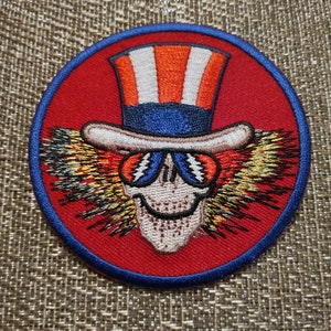 May include: Red, white, and blue embroidered patch of a skull wearing a top hat with a red, white, and blue band. The skull has a large smile and is wearing sunglasses. The skull is surrounded by a yellow, orange, and black flame-like design.