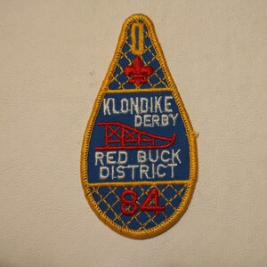 May include: A teardrop-shaped embroidered patch with a blue background and gold trim. The patch features the text "KLONDIKE DERBY RED BUCK DISTRICT 84" in white and red. A red sled graphic is also present.