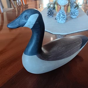 May include: A wooden goose figurine with a black head and neck, brown back, and cream-colored body. The goose is posed in a resting position, with its head held up. The figurine is on a dark brown wooden surface.