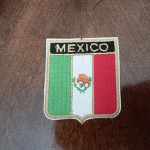 May include: A shield-shaped embroidered patch featuring the flag of Mexico with vertical green, white, and red stripes. The word MEXICO is stitched in gold thread on a black horizontal bar at the top. A small eagle emblem is centered in the white stripe.