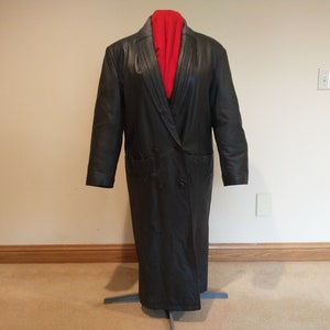May include: A long black leather coat with a double breasted closure and two pockets. The coat is on a mannequin and has a red scarf draped over the collar.