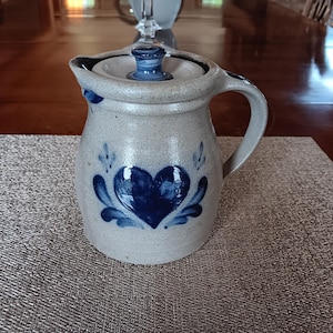 May include: A stoneware pitcher with a lid, featuring a blue heart design with leaf accents. The pitcher has a handle and spout, and is light gray in color. The pitcher is sitting on a textured placemat.
