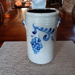 May include: A white ceramic crock with blue painted grape design and two handles. The crock is on a beige placemat.