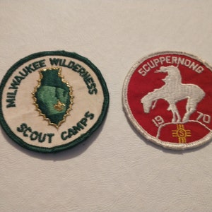 May include: Two vintage patches. One is round with "Milwaukee Wilderness Scout Camps" around a green arrowhead design. The other is red with a white rider on a horse and "Scuppernong 1970" text.