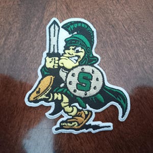 May include: An embroidered fabric patch featuring a cartoon Spartan warrior character in green and tan colors. The figure wears a helmet, holds a sword upright, and carries a round shield with a prominent green letter S in the center.
