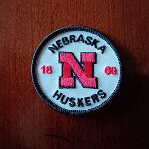 May include: Circular patch with a white background, featuring a red "N" and the words "NEBRASKA HUSKERS". The patch includes the numbers "18" and "69" in red. The border is black.