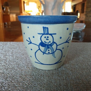 May include: A blue and white ceramic pot with a snowman design. The snowman is wearing a blue hat and scarf and has a blue button nose. The pot is decorated with blue dots.