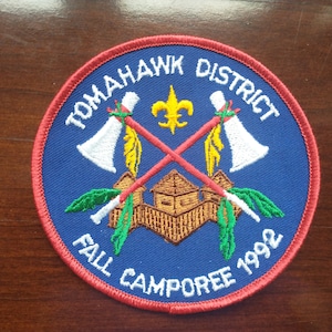 May include: Embroidered patch with the text "TOMAHAWK DISTRICT FALL CAMPOREE 1992". The patch features crossed tomahawks, a fleur-de-lis, and a stylized building, all set against a blue background with a red border.