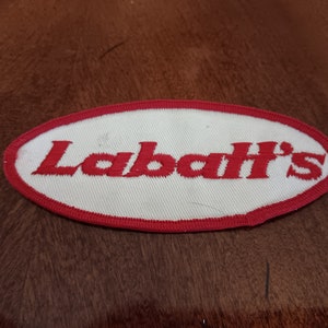 May include: An oval patch with the word "Labatt's" embroidered in red on a white background. The patch has a red border and is likely a logo or emblem.