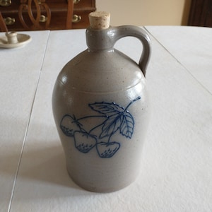May include: A gray stoneware crock with a blue and white design of strawberries and leaves. The crock has a cork stopper and a handle.