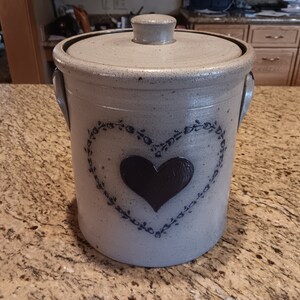 May include: A white ceramic crock with a lid, featuring a blue heart design with a black center. The crock has two handles and is decorated with a blue floral pattern.