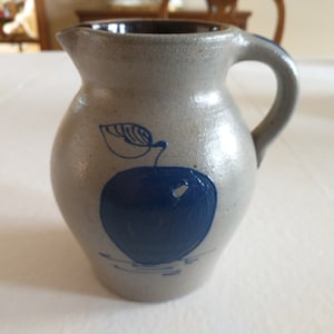 May include: A small gray ceramic pitcher with a blue apple design. The pitcher has a handle and a spout.