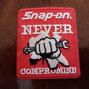 May include: Red square patch with white embroidered text and a graphic of a fist holding a wrench. The text reads "Snap-on, NEVER COMPROMISE". The patch has a white border.