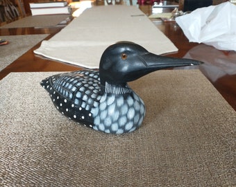 May include: A black and white wooden loon figurine with a black beak and an orange eye. The figurine is sitting on a brown woven mat.