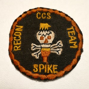May include: Embroidered patch with a black background and an orange border. The patch features a skull-like design with the words "CCS RECON TEAM SPIKE" in orange lettering. The design is centered on the patch.