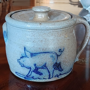 May include: A gray stoneware crock with a blue and white pig design. The crock has a lid and a handle.