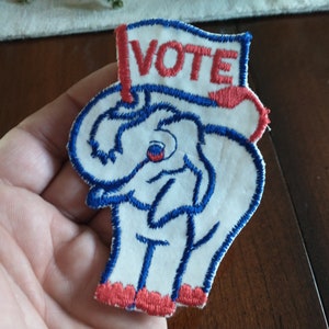 May include: A white fabric patch with a blue embroidered elephant wearing a red and blue hat with the word "VOTE" embroidered on it.