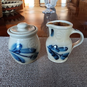 May include: A stoneware sugar bowl with a lid and a small pitcher. Both pieces are off-white with blue leaf designs and a stamped emblem. The sugar bowl is round, and the pitcher has a handle and spout. They sit on a textured placemat.