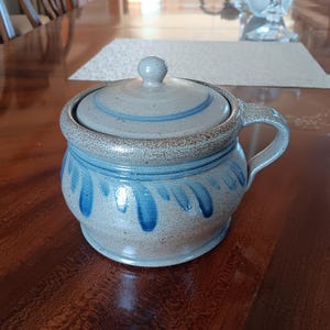 May include: A ceramic lidded jar with a handle, featuring a light blue and brown color scheme. The jar has a rounded shape with blue teardrop-like patterns. The lid has a small knob and blue stripes. The jar is likely for kitchen or dining use.