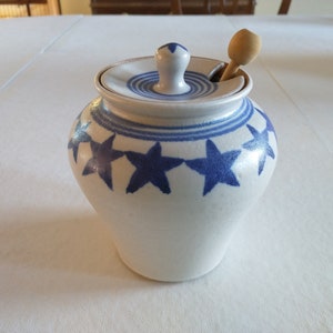 May include: A white ceramic honey pot with a blue star design and a wooden honey dipper.