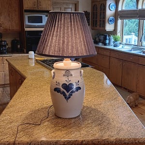 May include: A vintage-style lamp with a brown lampshade and a cream-colored base. The base is a stoneware crock with blue heart and floral designs. The lamp is on a granite countertop in a kitchen setting.