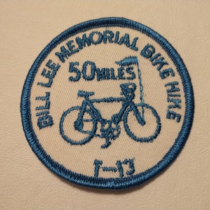 May include: A circular embroidered patch with a cream-colored background and a blue stitched border. The design features a blue bicycle graphic, a flag, and the text "BILL LEE MEMORIAL BIKE HIKE", "50 MILES", and "7-13" in blue embroidery.