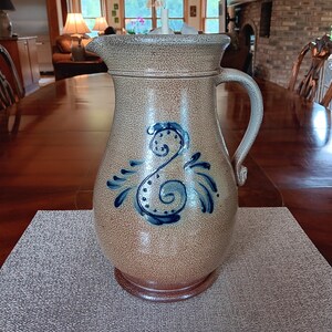May include: A stoneware pitcher with a speckled tan finish and a dark blue hand-painted swirl design on the front. The pitcher features a curved handle, a wide pouring spout, and a slightly darker brown base, showcasing a classic pottery form.