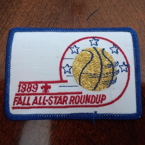 May include: A rectangular embroidered patch with a white background and a blue border. It features a gold metallic basketball, blue stars, and red text that reads 1989 FALL ALL-STAR ROUNDUP, along with a small red fleur-de-lis emblem.