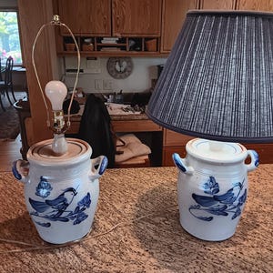 May include: A ceramic lamp base with a blue bird and floral design, paired with a navy blue lampshade. The lamp base has two blue handles and a light bulb. The lamp is on a brown surface.