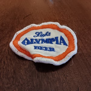 May include: Embroidered patch with the words "Light Olympia Beer" in blue lettering on a white background. The patch has an orange border and a white stitched edge. The patch is slightly wrinkled.