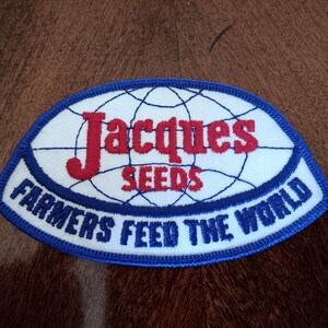 May include: An oval-shaped patch with a white background and a blue border. The patch features the words "Jacques SEEDS" in red and "FARMERS FEED THE WORLD" in blue, with a globe design.