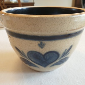 May include: A cream-colored ceramic bowl with a blue heart design and blue trim. The bowl is glazed and has a rustic look.