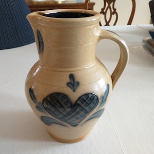 May include: A brown ceramic pitcher with a blue heart design and blue floral accents. The pitcher has a wide mouth and a handle.