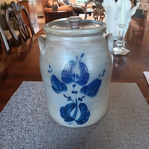 May include: A vintage stoneware crock with a lid, featuring a blue floral design. The crock has two handles and is set on a gray placemat. The text "Winona Pottery" is visible on the crock.