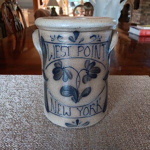 May include: A tan stoneware crock with two handles and a wide rim. The crock features blue lettering that reads "WEST POINT NEW YORK" with floral designs. The antique-style container is likely used for storage.